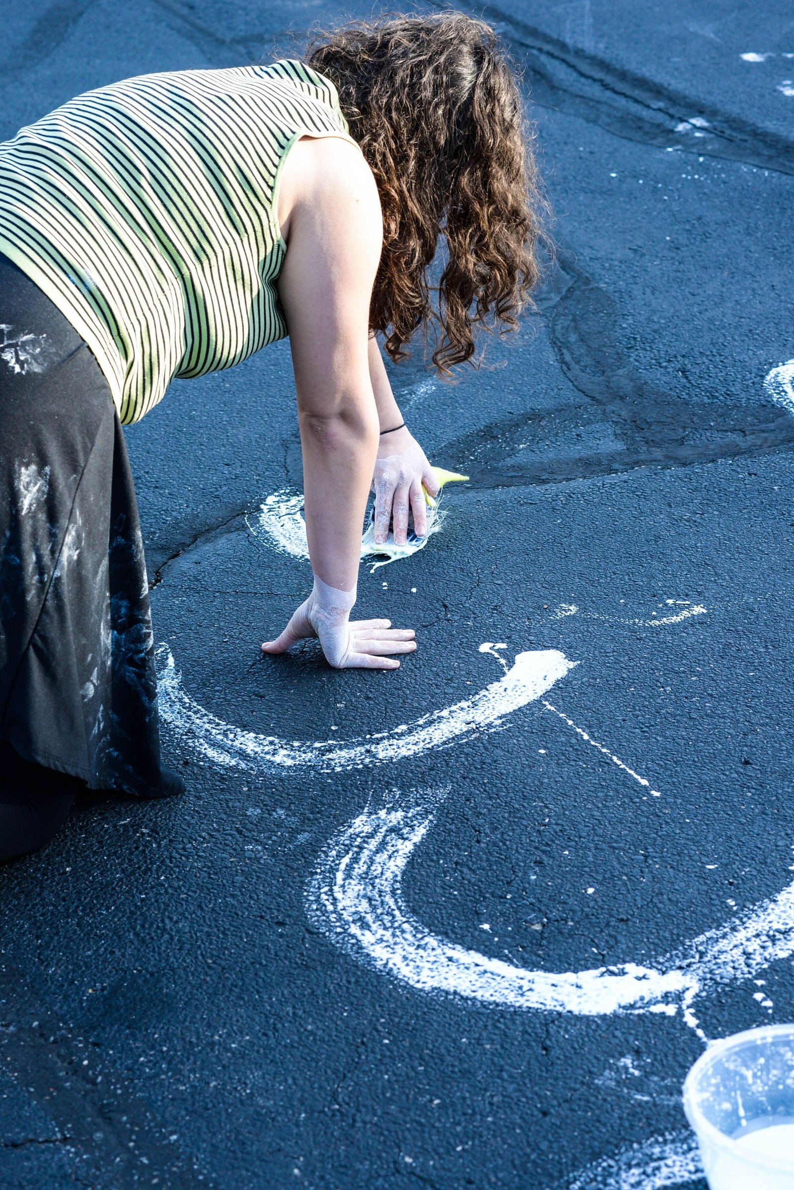 A person of light skin tone paints with their hands on dark asphalt. They are close to the ground as they make their art.