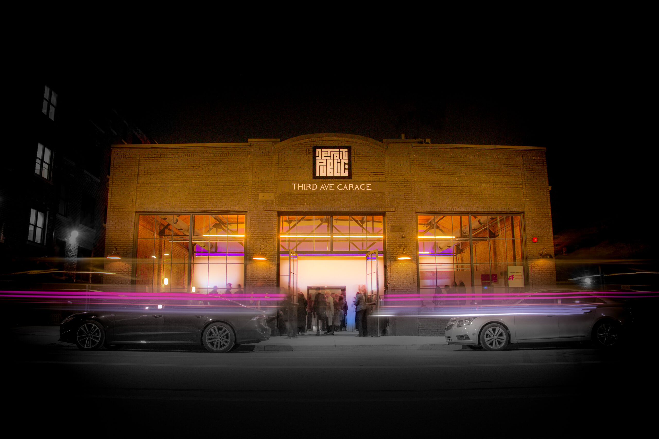 A facade of a one-story beige-colored building with lettering that reads "Third Ave Garage" under a stylistic logo of the Detroit Public Theatre. The photo was taken during night time, so lights are shining through the building's large square windows, and cars are trailing the foreground of the image.