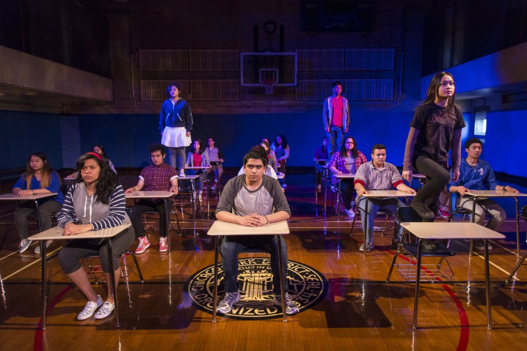 Around a dozen young actors in a classroom set during a theater performance. Some are sitting at their desks, while others stand on the table. There is blue light shining in the background.