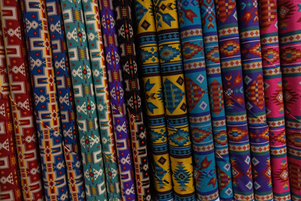 A close-up of a stack of fabric bolts on a shelf. They range from reds, blues, greens, yellow and black, and have Native American-inspired patterns for regalia-making.