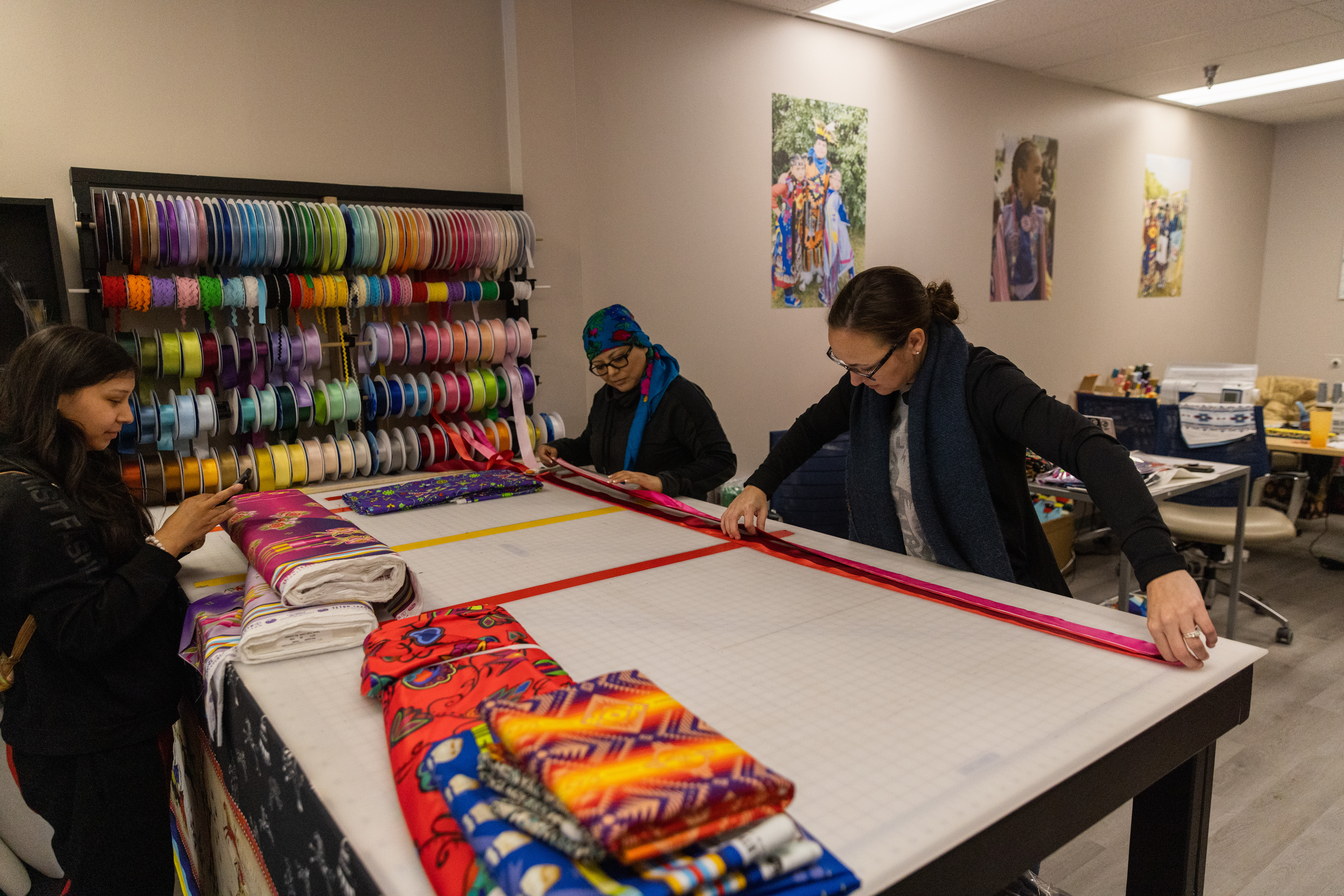 Three people stand around a cutting table flanked by colorful bolts of fabrics and rolls of ribbons. Two of them are on one side of the table gathering ribbons together as they fulfill an order for the customer on the other side of the table. The customer is waiting, while looking at their mobile phone.