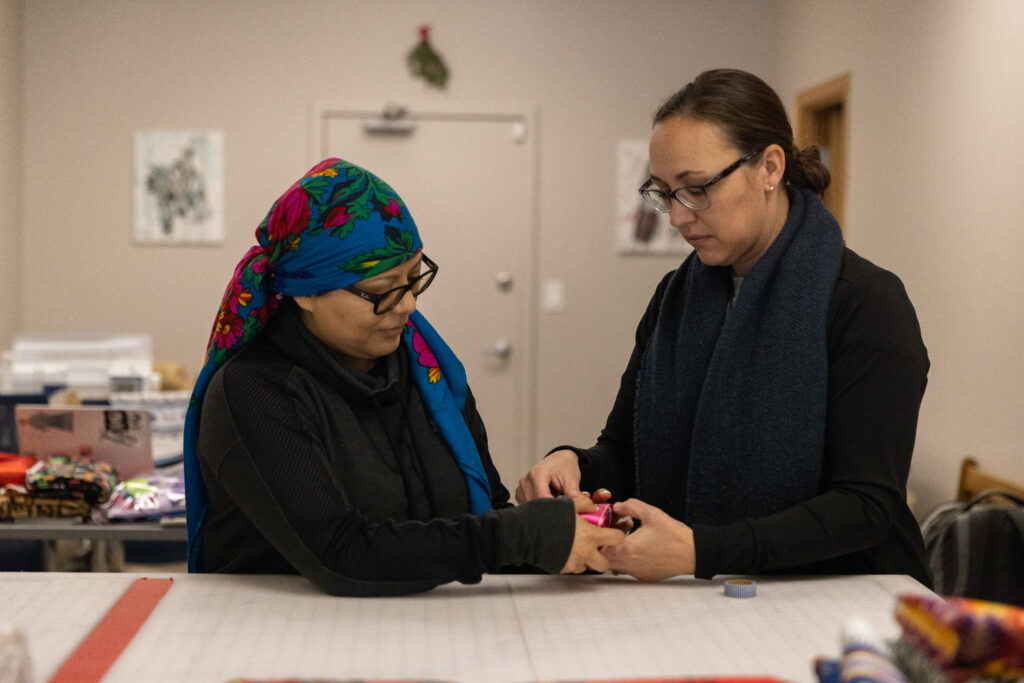 Two people of medium light skin tone hold a stack of ribbons together on a cutting table. Both are wearing glasses and dark colored clothing. One of them is wearing a bright blue floral headscarf and the other has a thick navy scarf around their neck.