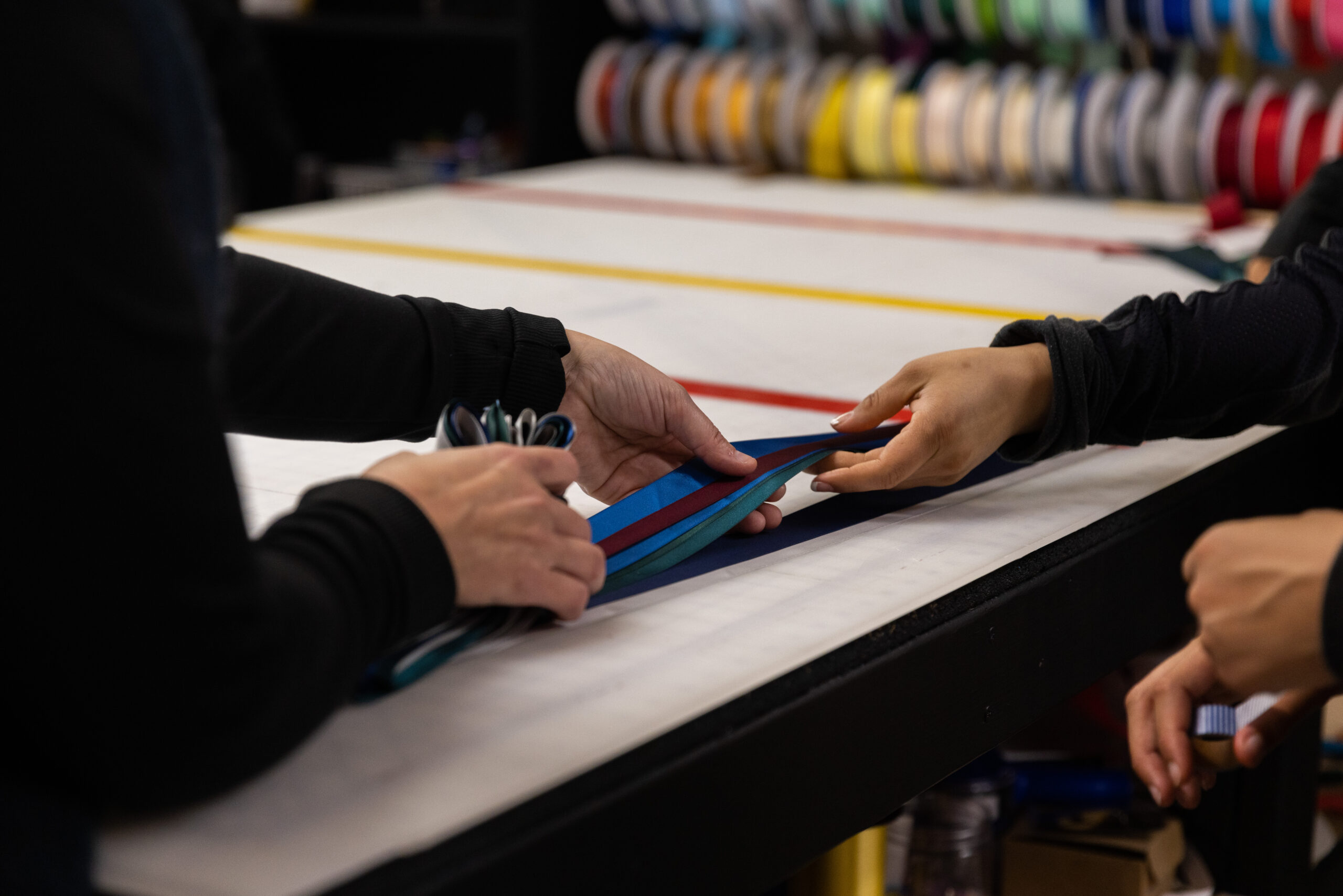 A person folds a collection of ribbons, in hues of blues, greens and deep red, on top of a cutting table. Their hands are in focus in the photo as another set of hands helps them with the task. There are a set of ribbon spools far in the background.