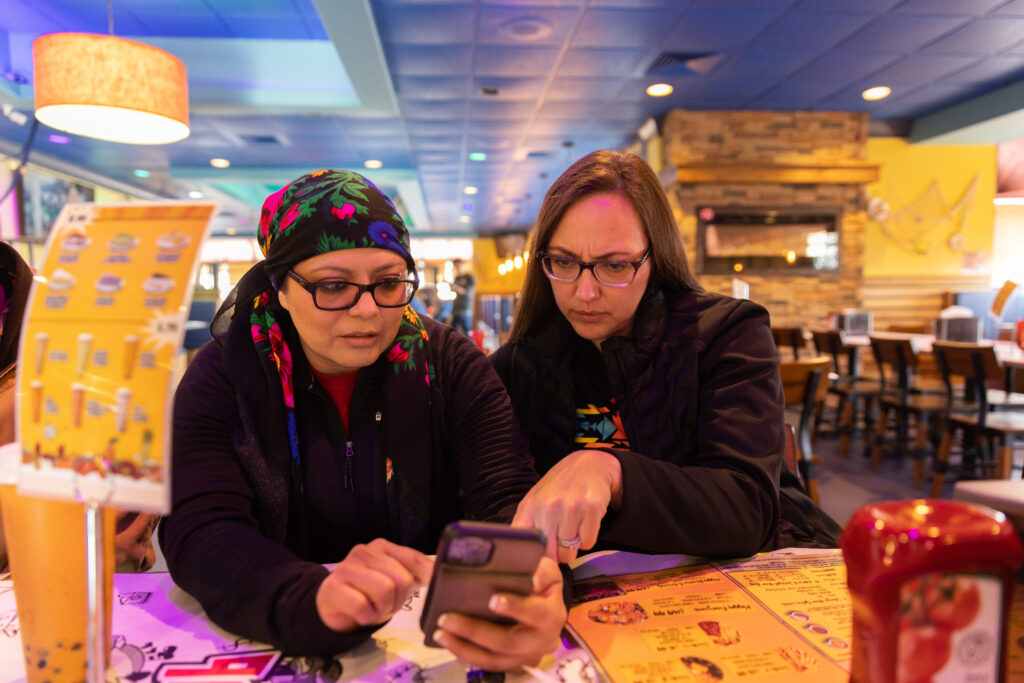 Two people of medium light skin tone sit at a restaurant table, looking and gesturing to something on a mobile phone. Both of them are wearing dark-framed glasses and dark-colored clothing; one of them is wearing a black floral headscarf. The restaurant feels warm with yellow lighting.