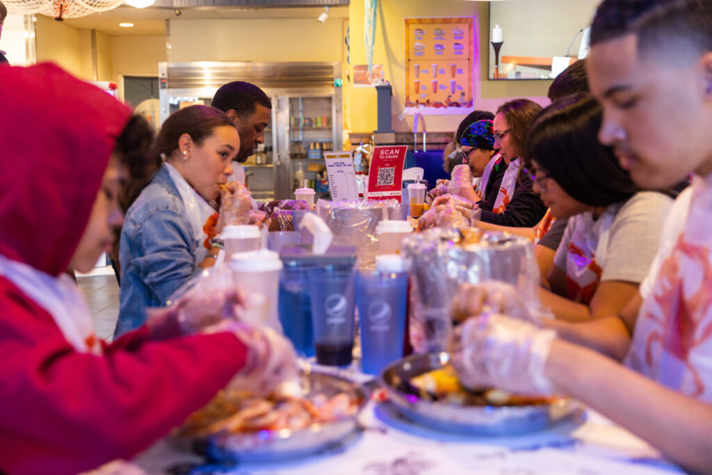 A family of young kids, teenagers and adults sit at a long table as they share a meal together at a restaurant. They are wearing light plastic gloves as their enjoy seafood broil.