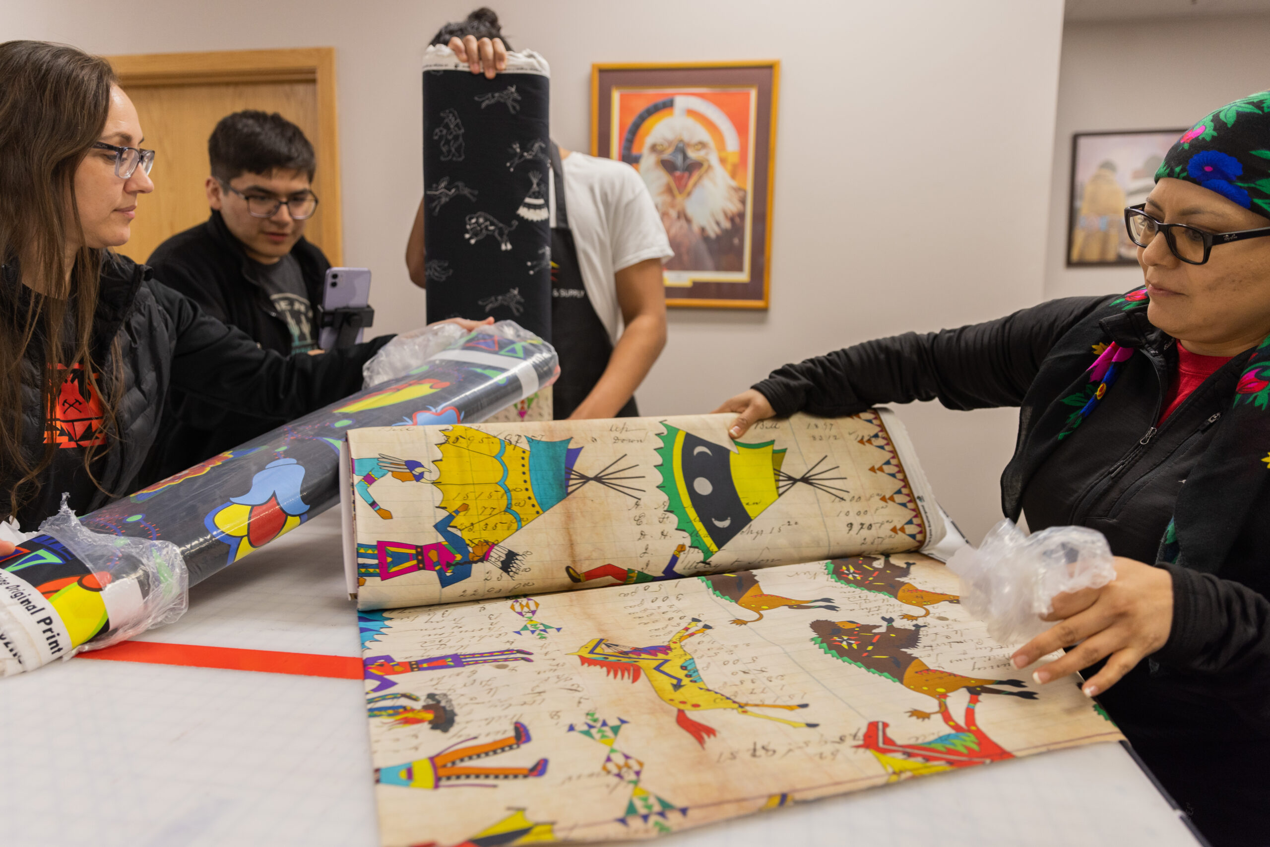 Two people of medium light skin tone unpack bolt of fabrics as two teenagers look on and help them.