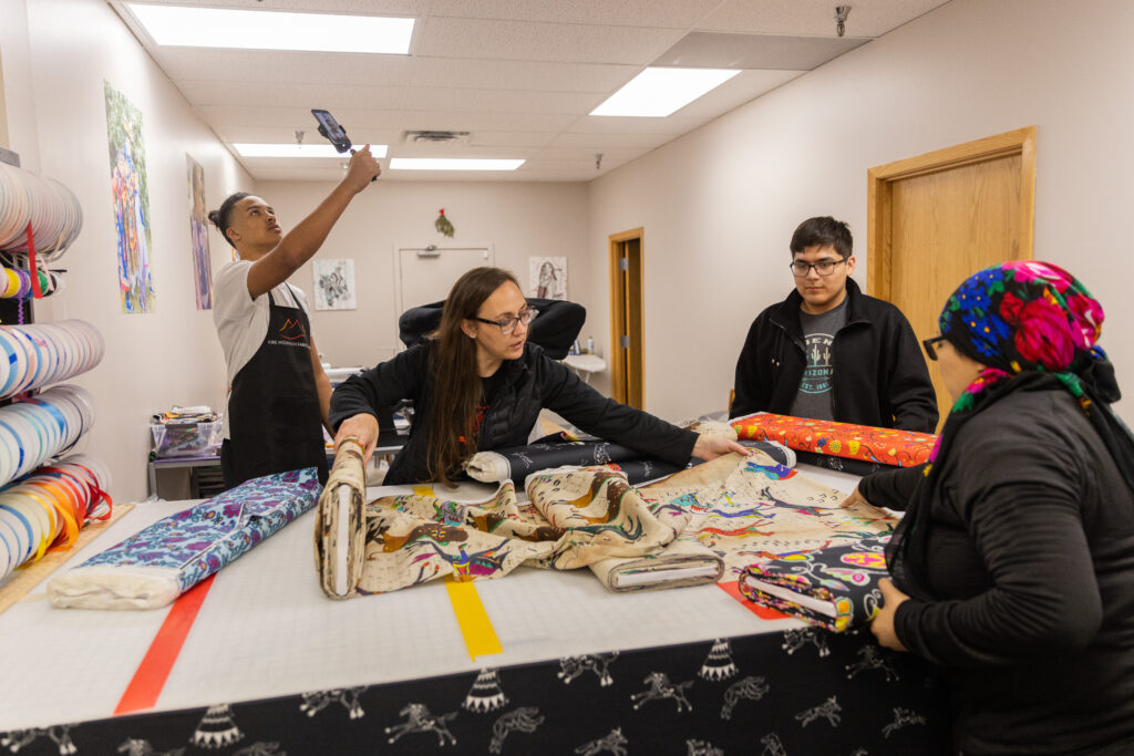 Two people handle and unroll bolts of fabric on a cutting table as one teenager looks on, and another holds up a phone on a handheld tripod. The fabrics are colorful and patterned.