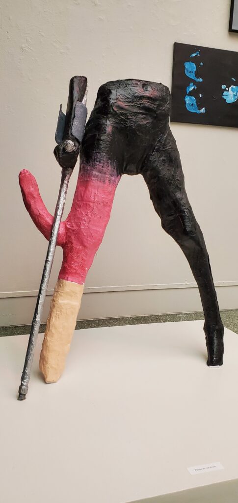 A sculpture of the bottom half of a person. One side is painted black, and the other leg is red on top and beige at the bottom. A offshoot of the leg holds up a cane.
