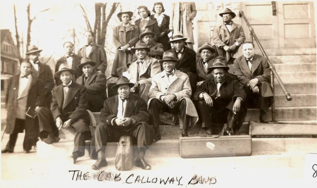 An archival black and white photo of a group of people dressed to the nines sitting on the steps of a building. Most of them are wearing hats and long wool coats.