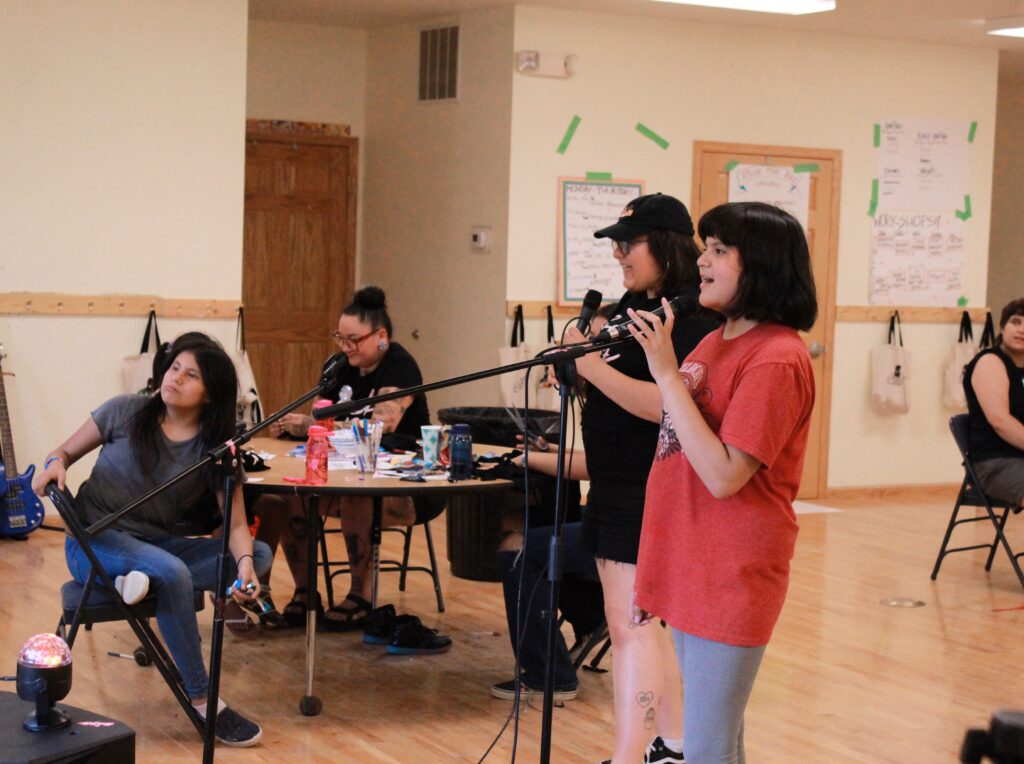 A young person sings into a microphone accompanied by an adult, while others look on and listen to them.