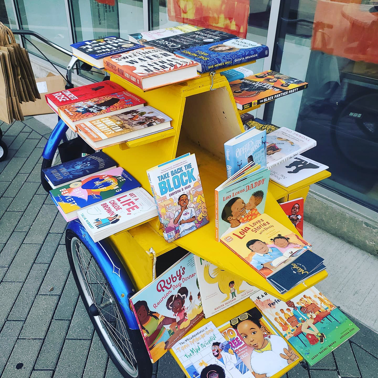 A collection of books rest on a handmade, rigged bike shelf for a mobile bookstore. The yellow shelf rests on the back of a blue three-wheeler bicycle.