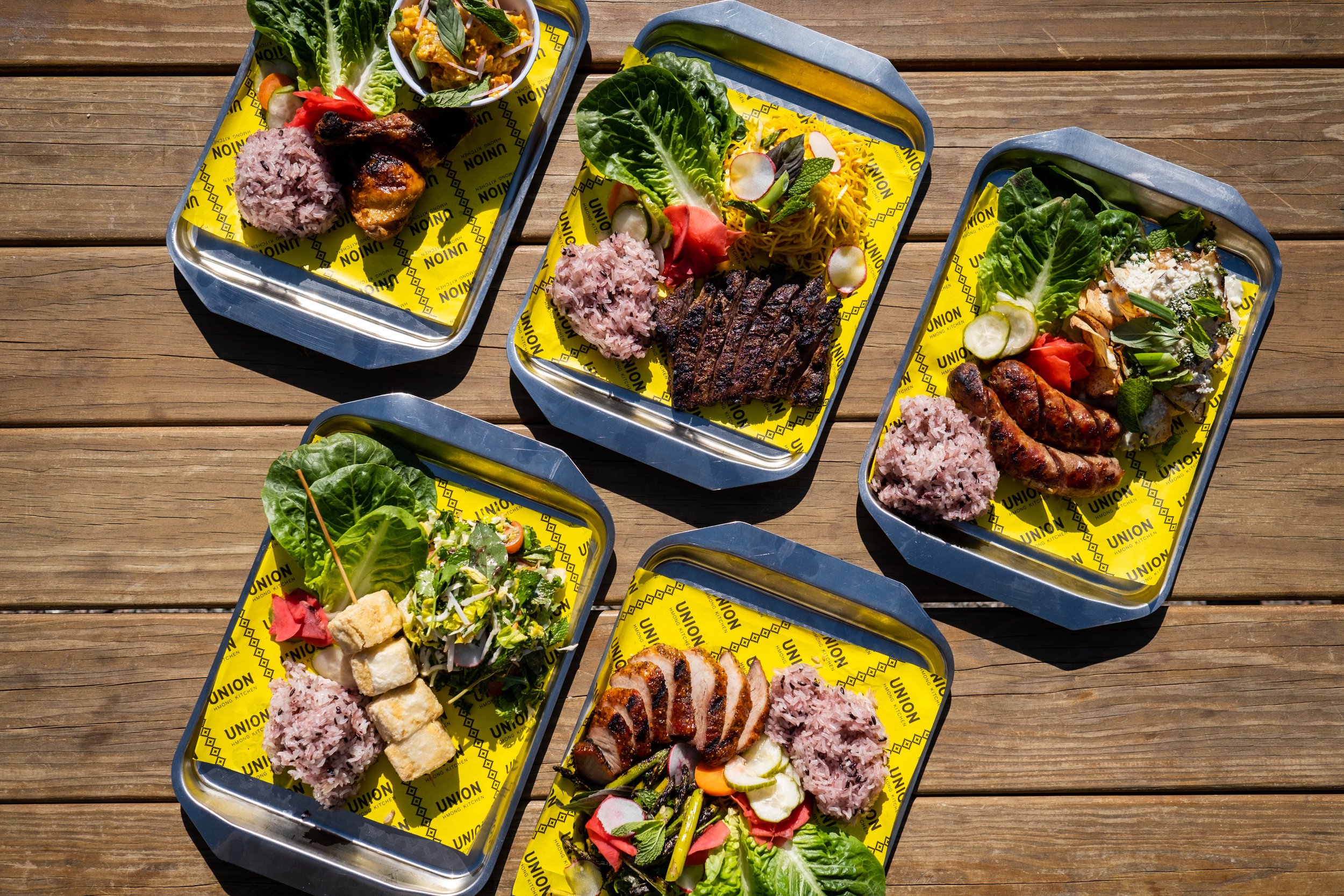 Five rectangular silver trays showing foods from the menu of Union Hmong Kitchen in Minneapolis, MN. The trays carry a variety of meats, purple sticky rice and some vegetables.