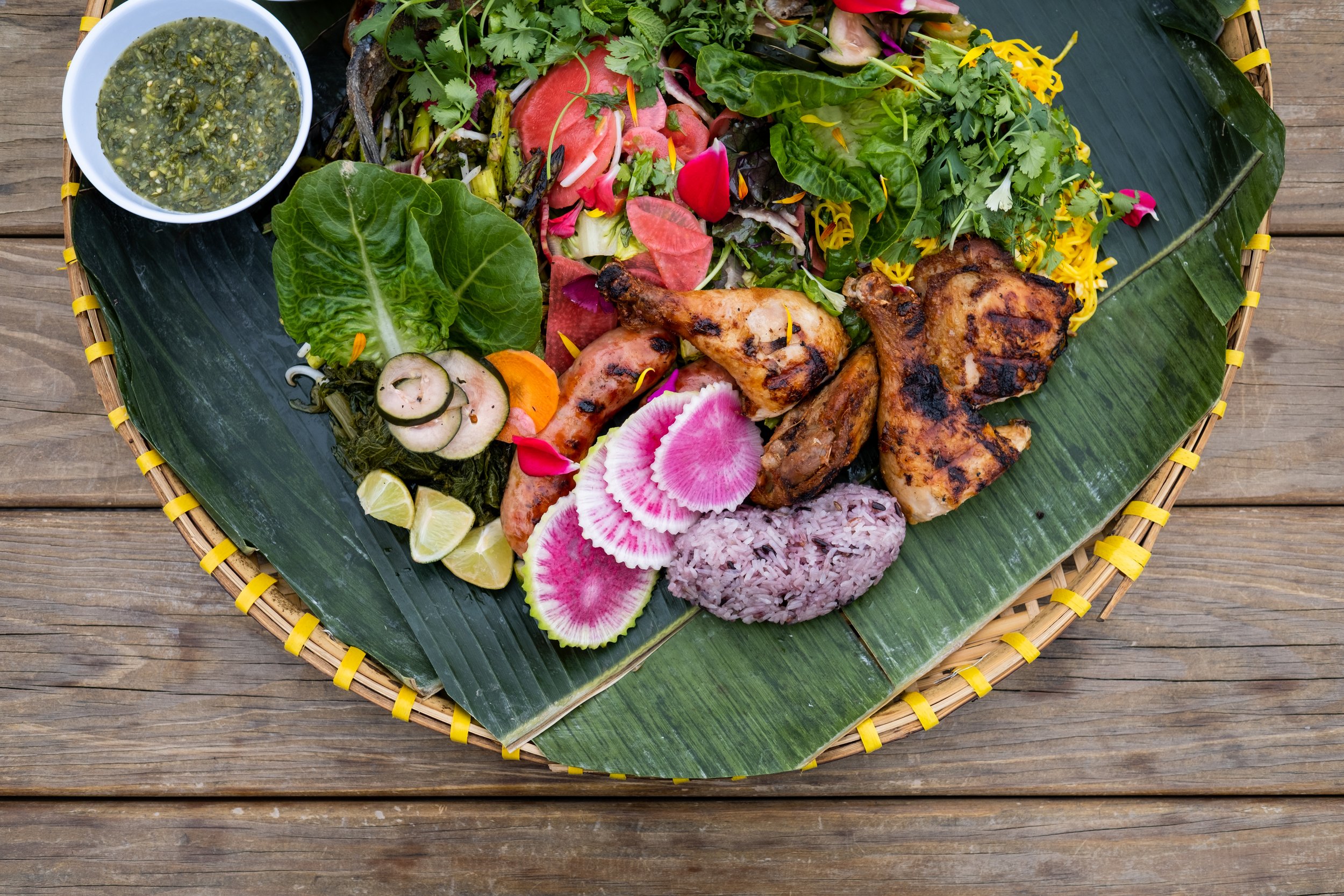 A circular woven bamboo tray lined with banana leaves holds a colorful variety of meats and vegetables as well as purple sticky rice. It's a shared plate served at the Union Hmong Kitchen in Minneapolis, MN.