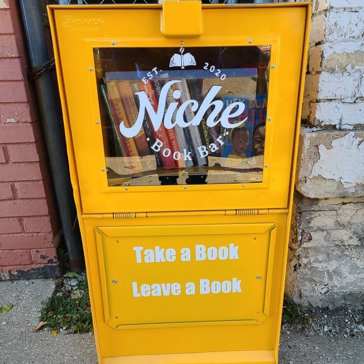 A yellow metal magazine stand turned into a free library. There's lettering on the box that reads "Est. 2020 Niche Book Bar" followed by "Take a book, leave a book."