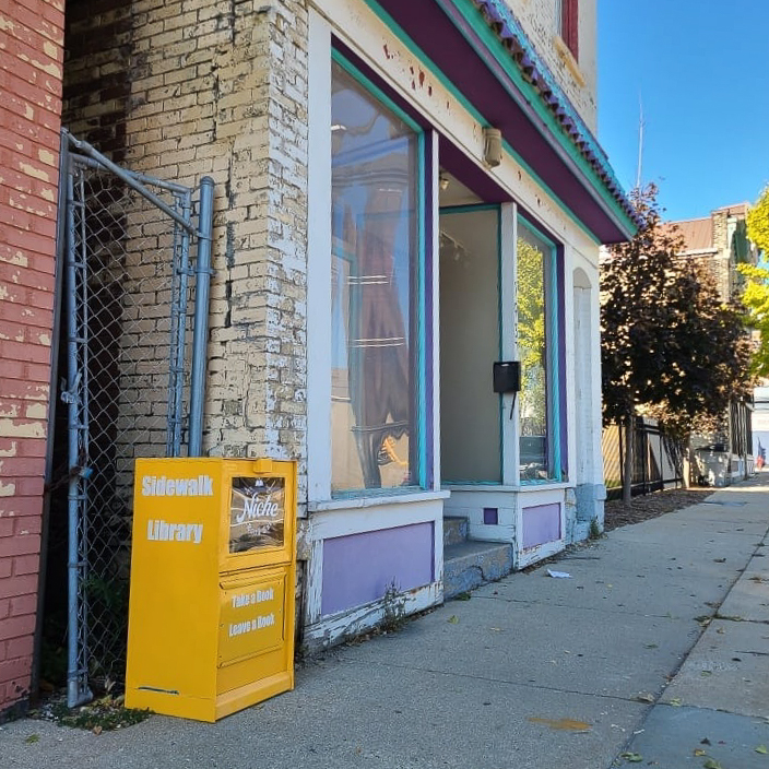A view of a street with a bright yellow newspaper stand turned into a free library. The building in view has large windows.