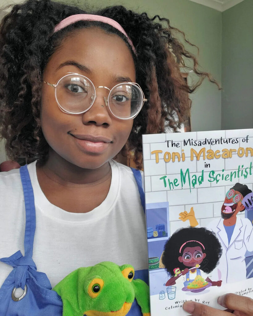 A person with dark skin tone holds up a book titled, The Misadventure of Toni Macaroni in The Mad Scientist. They are dressed like the main character of the book, wearing round glasses and are in a white t-shirt and overalls.