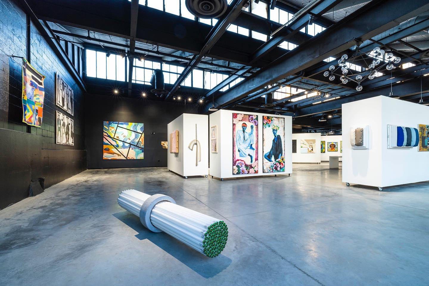 A wide shot of a gallery floor showcasing a mix of two-dimensional and three-dimensional artworks. Some of them are hanging on moveable white walls and other colorful works are on the black-colored walls of the space. In the foreground, there is a sculptural work made of fluorescent tube lights.