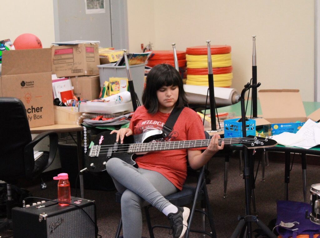 A young person wearing a red shirt and grey pants sits with their legs crossed, playing a bass guitar.