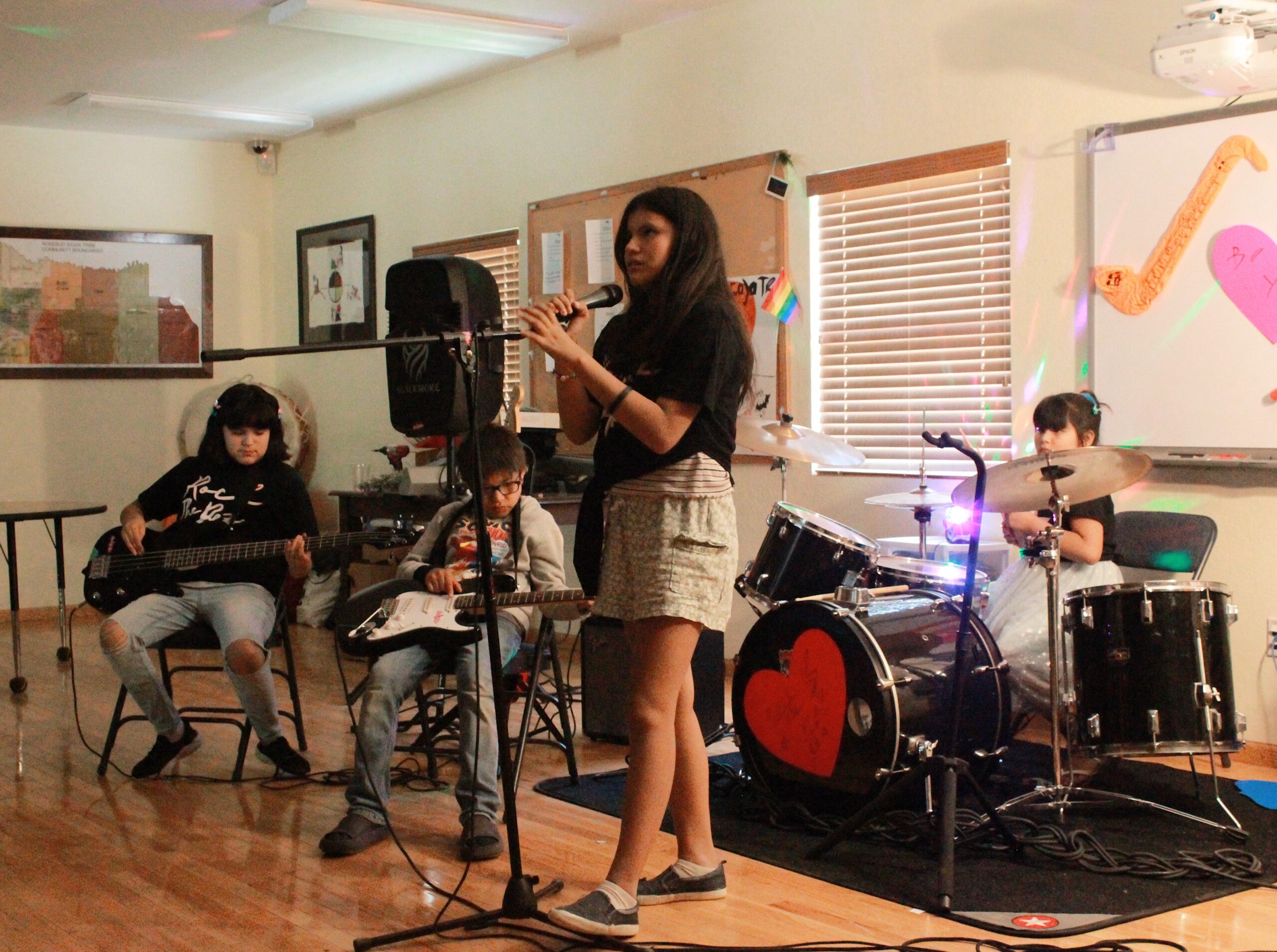 Four young people perform as musical band. One sings into a microphone, while the other three play the bass guitar, electric guitar, and drums.