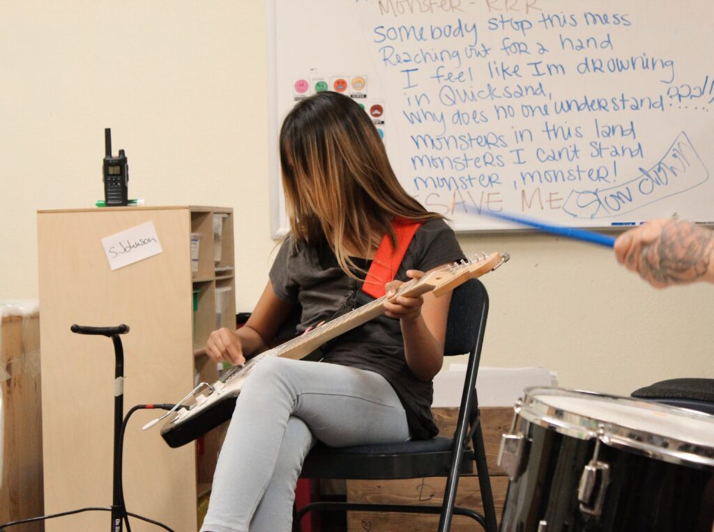 A young person sits and plays the electric guitar during a music camp.