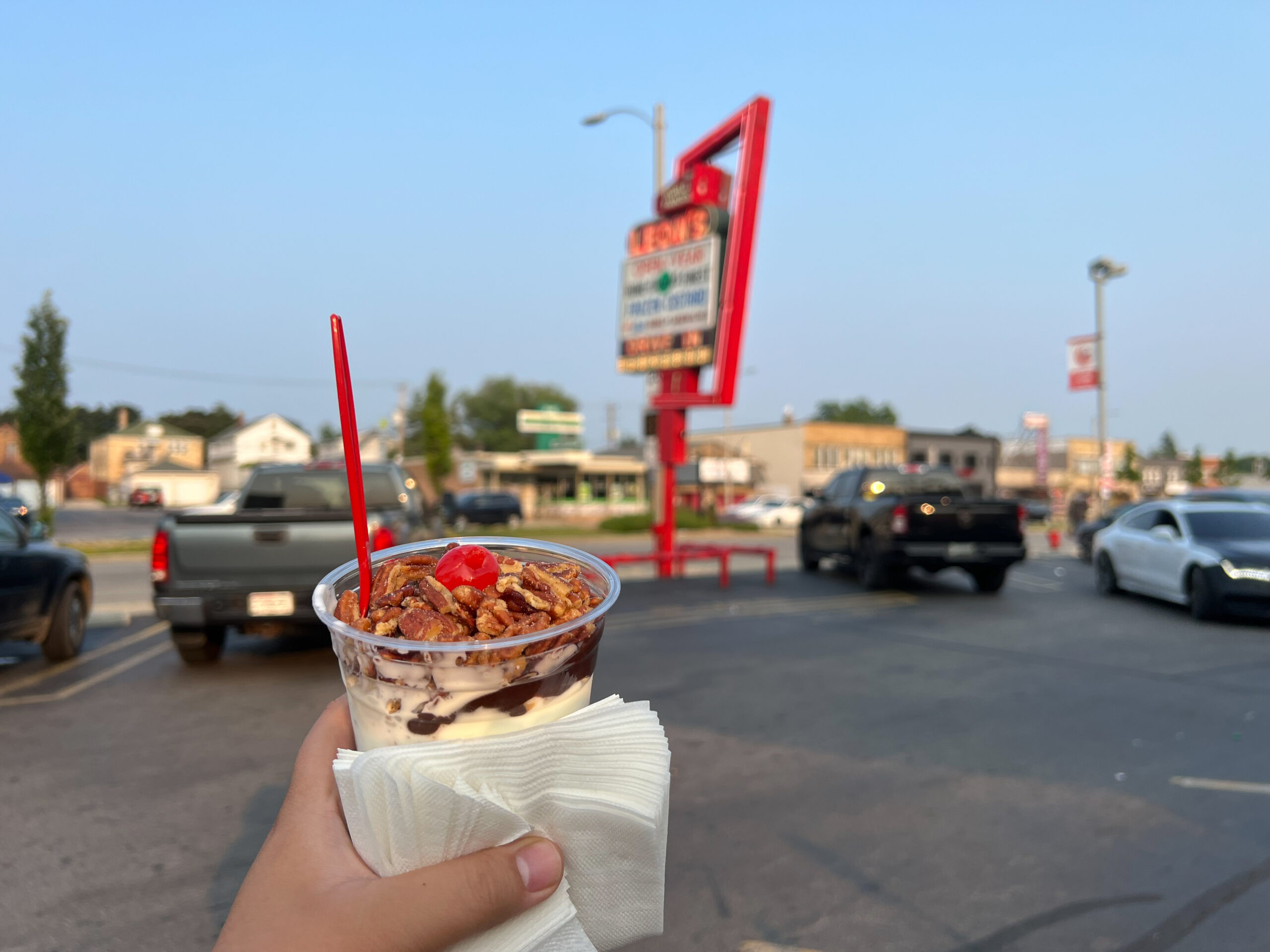 A hand holding up a plastic cup of custard with fudge, nuts, and a cherry, in front of a sign reading "Leon's Drive-in."