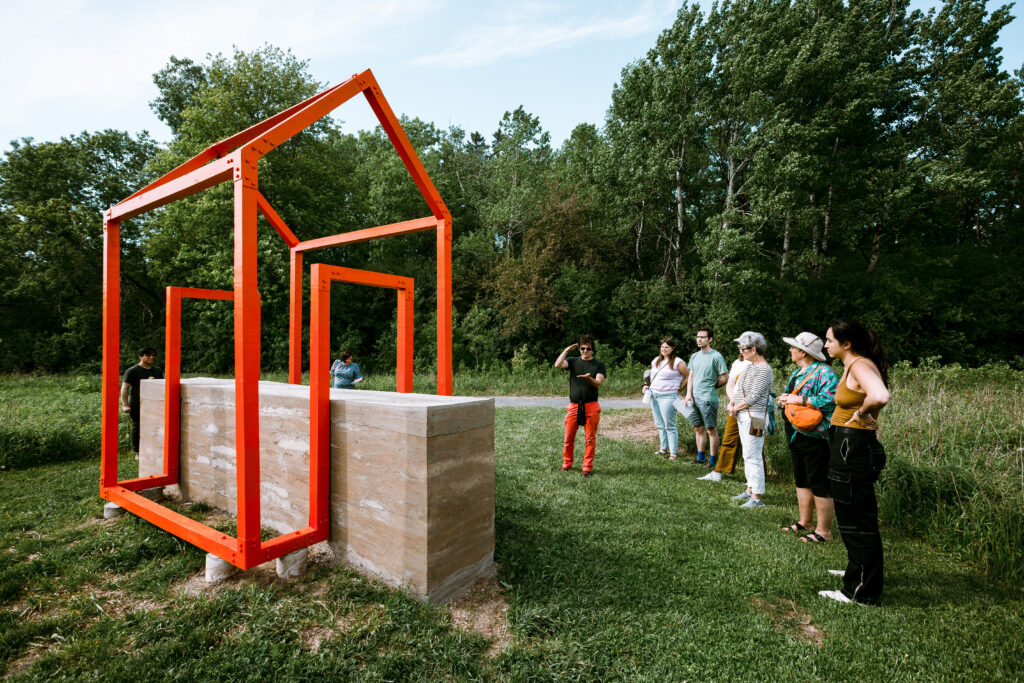 A sculpture of the outline of a orange house, with a slab of mud within it. A group of people look at an artist as he explains the work.