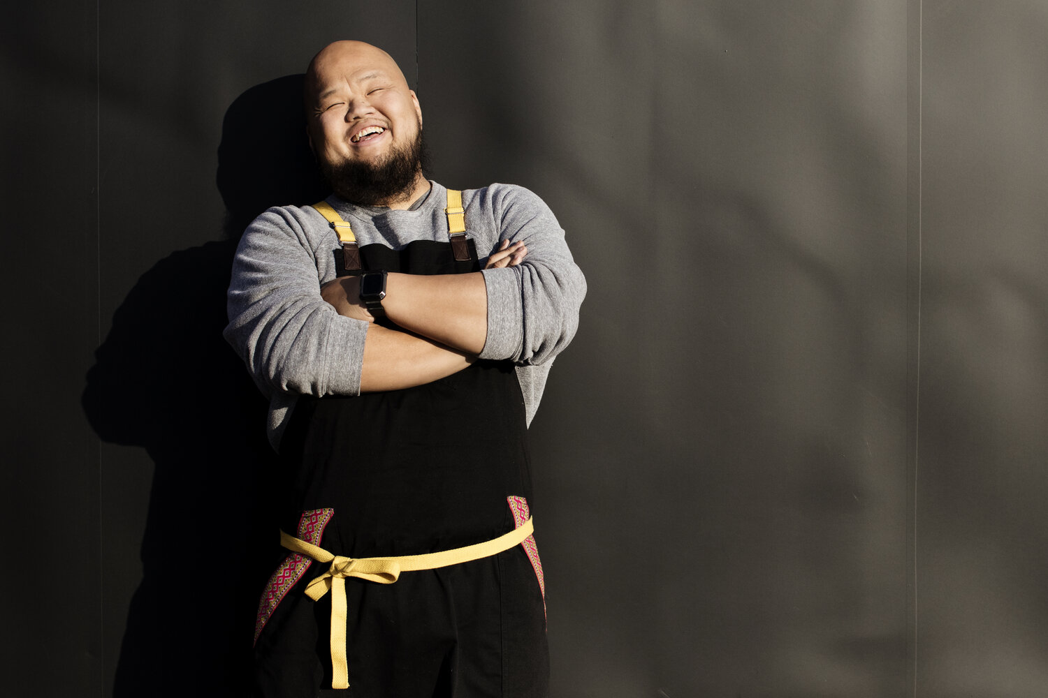 A person with medium light skin tone smiles as they cross their arms across their chest. They are wearing a grey sweatshirt under a black chef's apron. They have a dark beard and a shaved head.