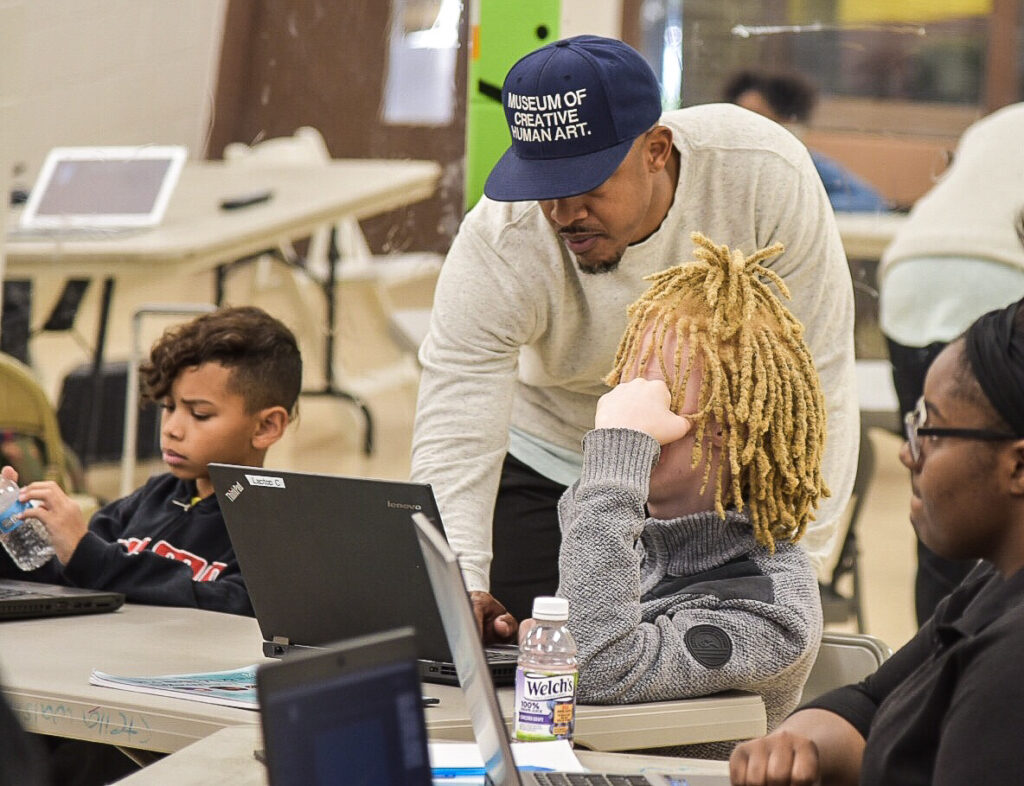 A person of medium dark skin tone hunches over to help a student with work on their laptop.