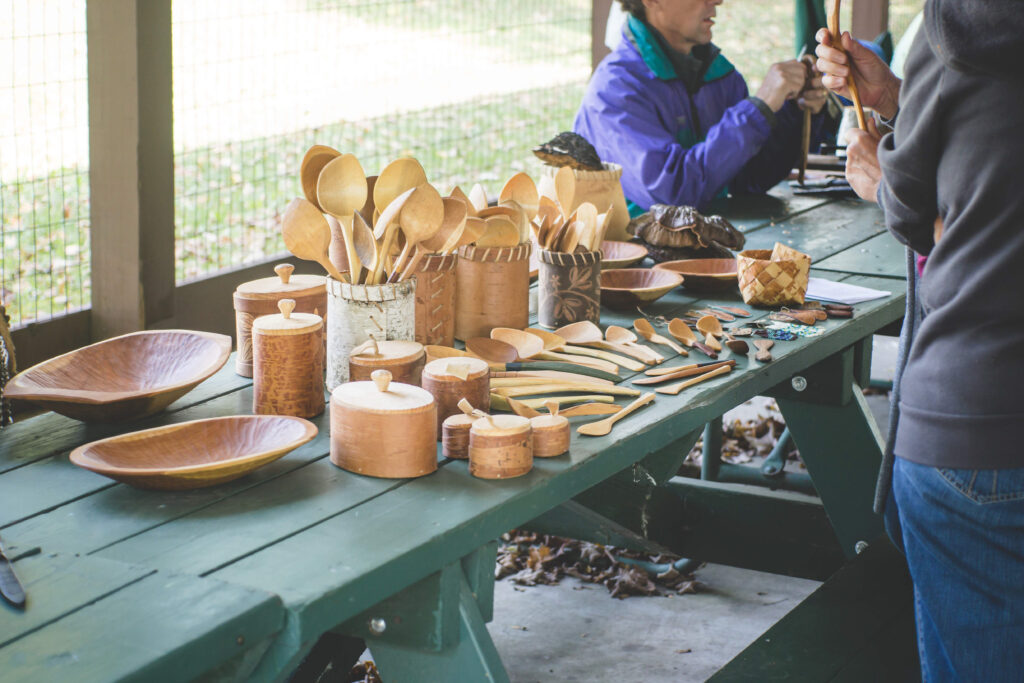 A variety of handmade wooden items are presented on a dark green picnic table. There are spoons of varying shapes and sizes, as well as lidded containers and bowls.