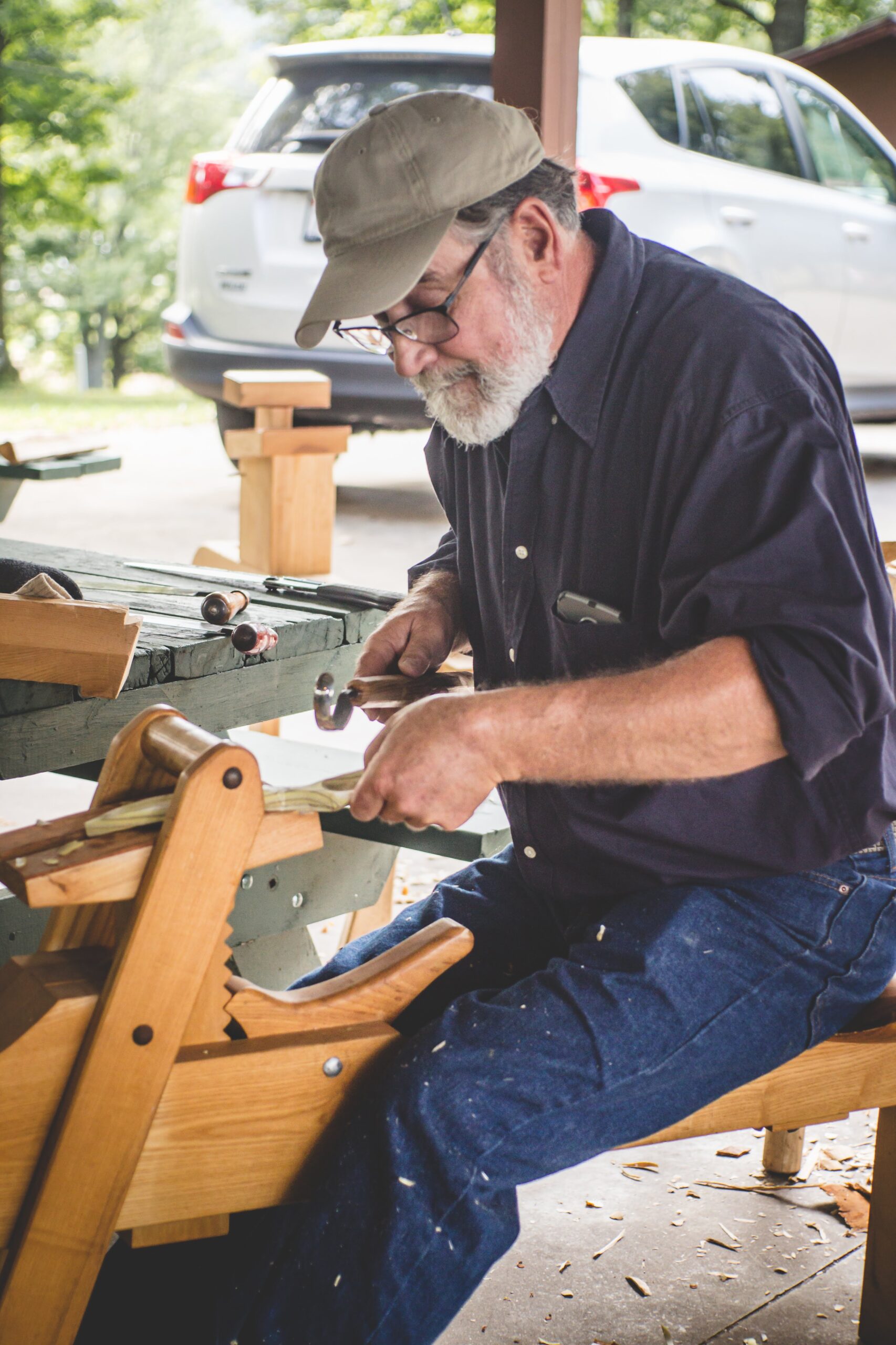 A bearded person of light skin tone sits on a woodworking bench called a shave horse as he carves a wooden spoon. He is wearing a beige baseball hat, glasses, a navy blue button-up shirt and blue denim jeans.