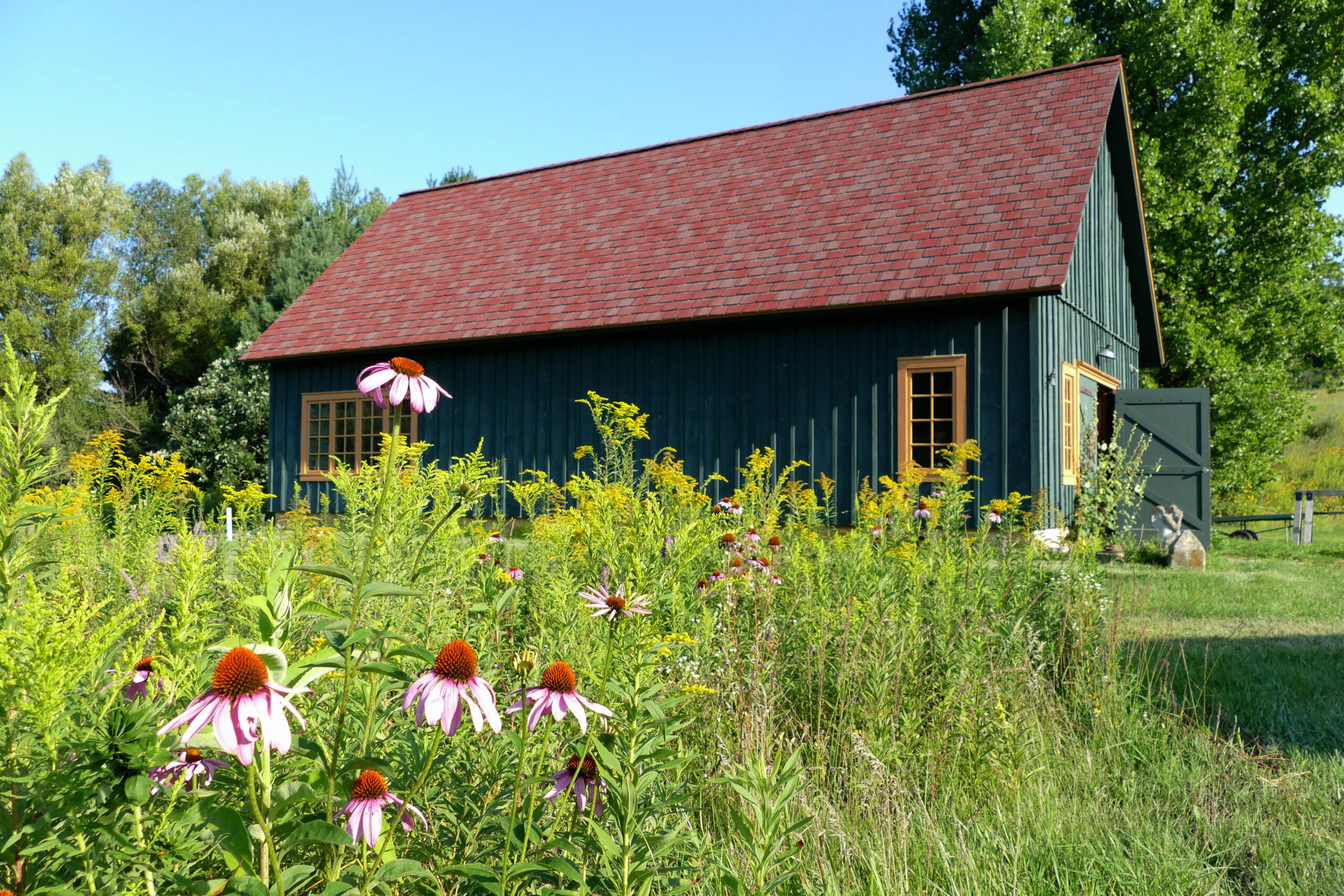 Perennial flowers, including coneflower and goldenrod, bloom beside a large dark green building used as an art studio and workshop. The building has several windows with mustard-colored trim and one of the large barn-door entrances is open.