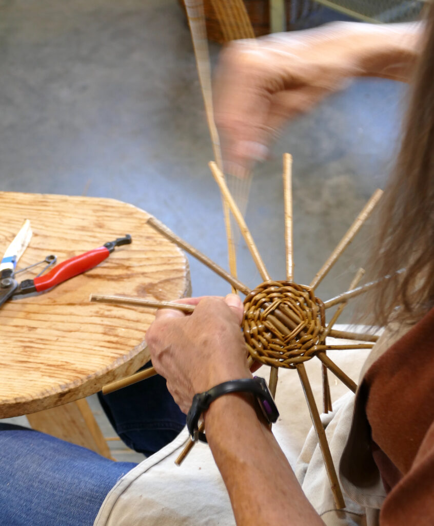A hand holds the start of a woven willow basket. There is a base structure made of criss-crossing sticks and woven on it with thinner branches is a concentric pattern.