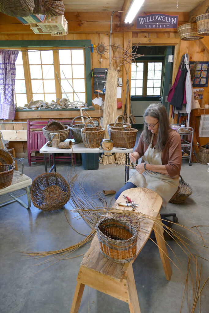A person of light skin tone and greying hair is in the middle of weaving a basket. She is surrounded by basket weaving materials and tools as well as a selection of wooden baskets of varying sizes and shapes.