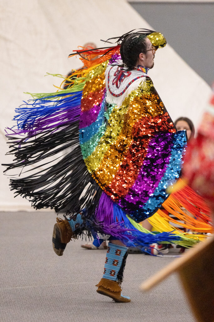 A person of light skin tone dances in their pride flag inspired sequined shawl.