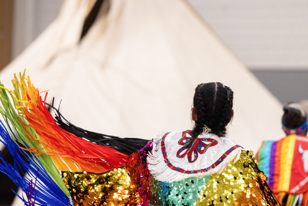 A person with dark hair faces away as their colorful sequined regalia with rainbow-colored fringe flails in the air.