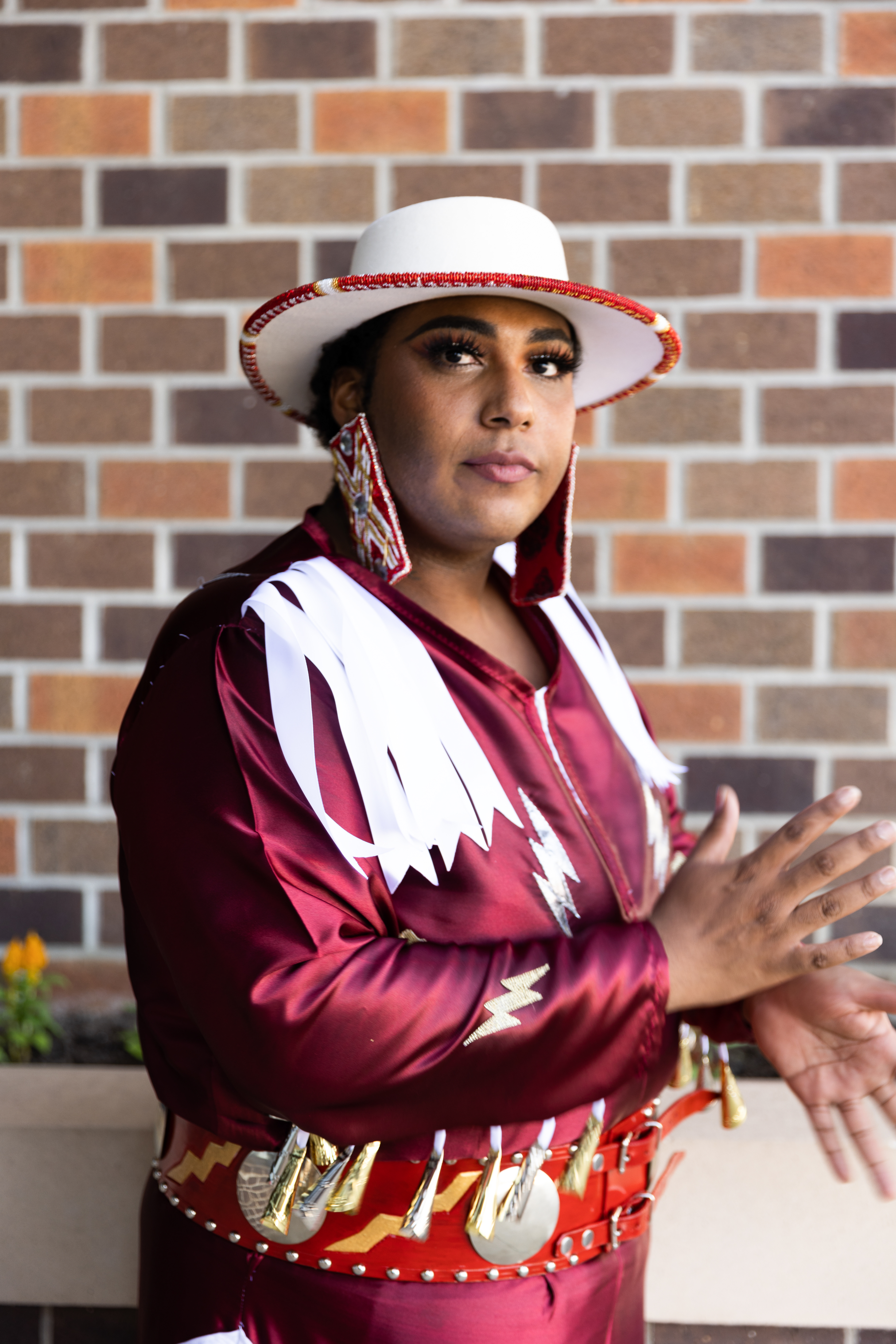 A person with medium-dark brown skin wearing large rectangular earrings, a white hat, and a red jacket with white fringe, looking at the camera.