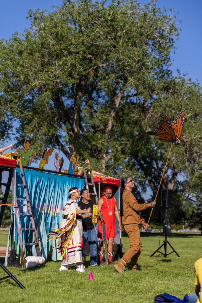 Four people in a variety of clothing during an outdoor theater performance. One person wearing an all-brown sweatsuit holds a enlarged depiction of a monarch butterfly, as the other three people look on.
