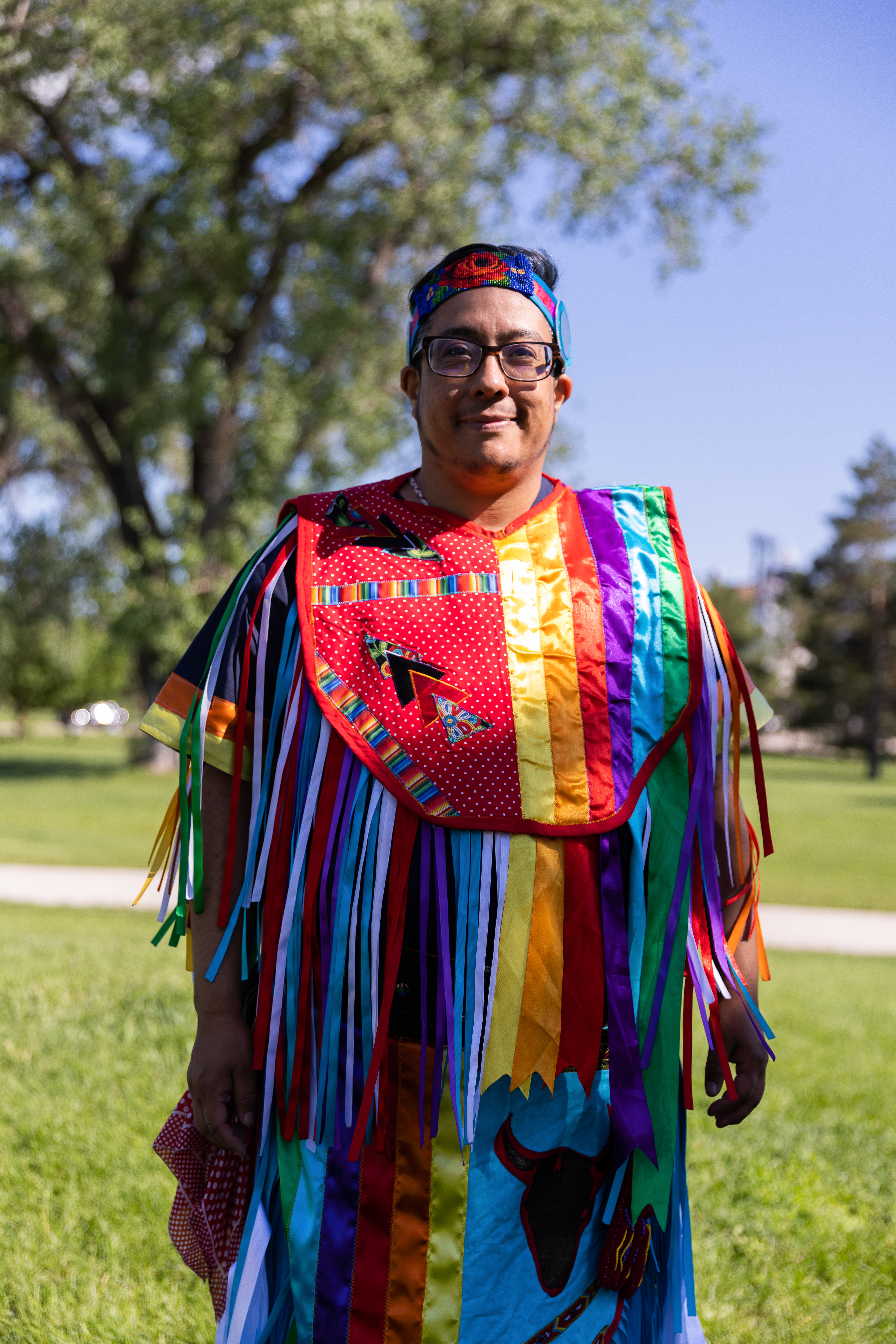 A person with medium-tone skin wearing colorful regalia and smiling at the camera.