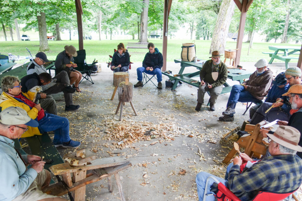 Over a dozen people sit in a big circle, and the floor is covered in wood chips and shavings. Most of the people are looking down at their hands as they hold a tool and carve a wooden spoon.