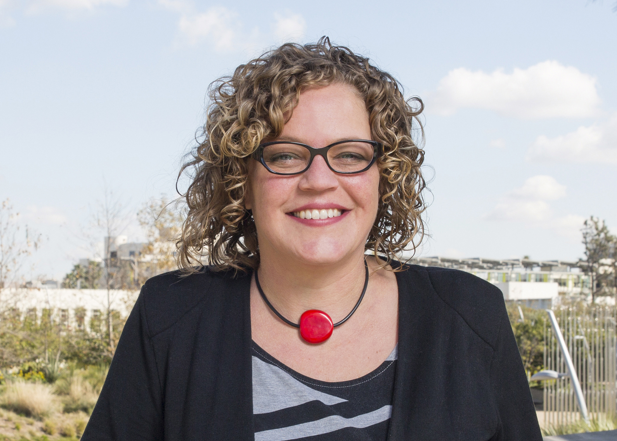 A person of light skin tone wearing dark-rimmed glasses, a decorative red necklace and a patterned top smiles as they look at the camera. They have curly light brown hair.