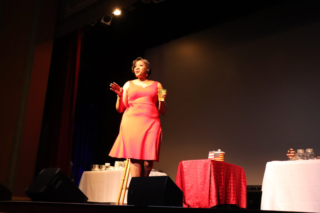 A woman with dark skin and a red dress speaks on stage.