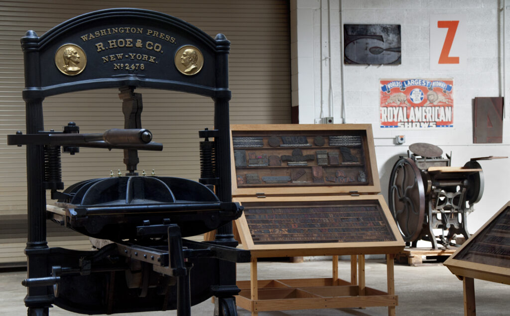 An old printing machinery sits next to a display of old wood type blocks in a museum space.