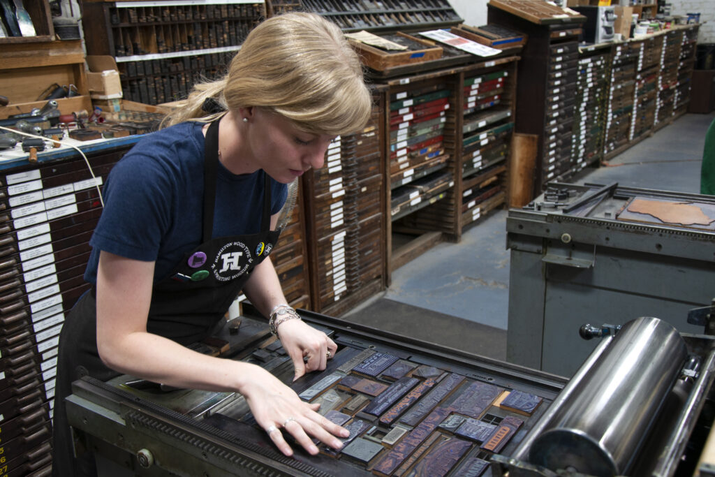 A person of light skin tone and light blonde hair bends over a printing press and arranges wood blocks.
