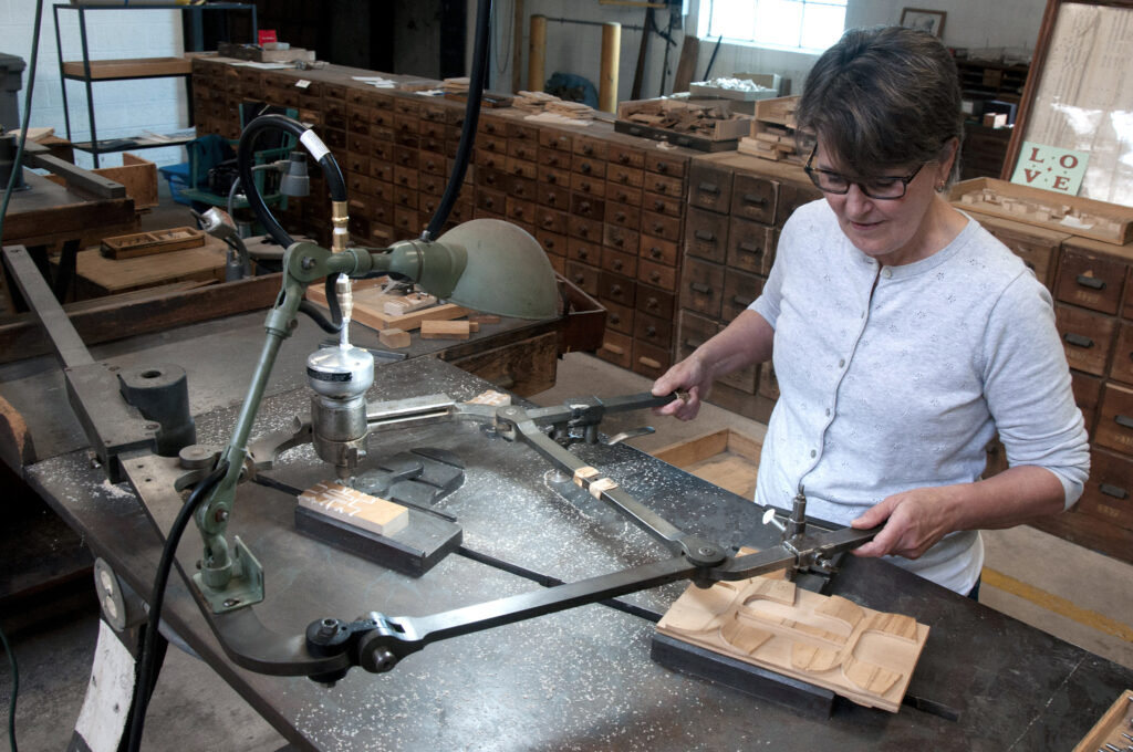 A person of light skin tone uses a metal machinery with extended arm-like levers to cut wood type.