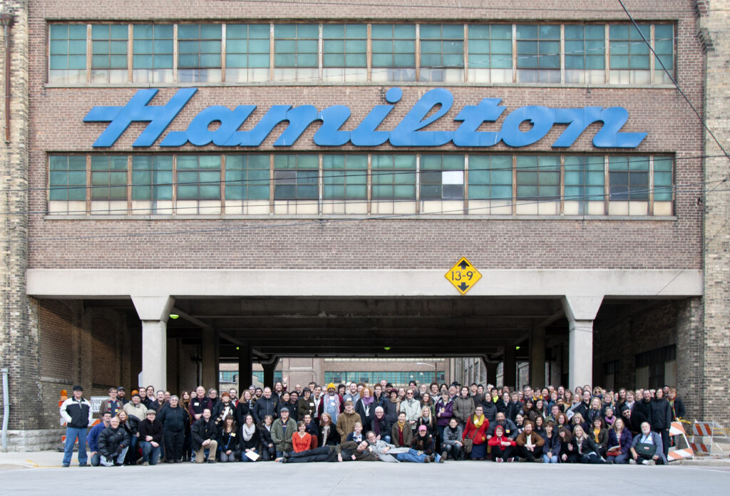 A large group of people pose under a large sign on a brick building that reads 'Hamilton' in blue cursive letters.