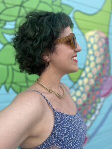 A side profile of a person with medium light skin tone with a big smile. They have dark, short curly hair with accents of deep green. They are wearing a large-framed, leopard-print sunglasses.
