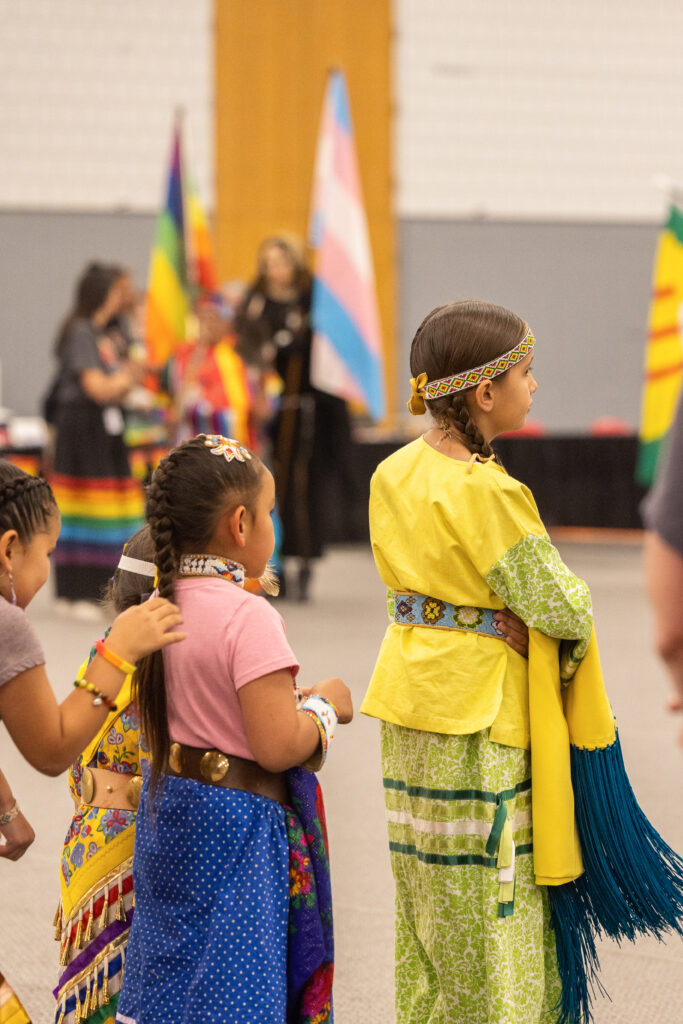 A group of young children line up during a powwow. They are all dressed in a variety of colorful regalia.