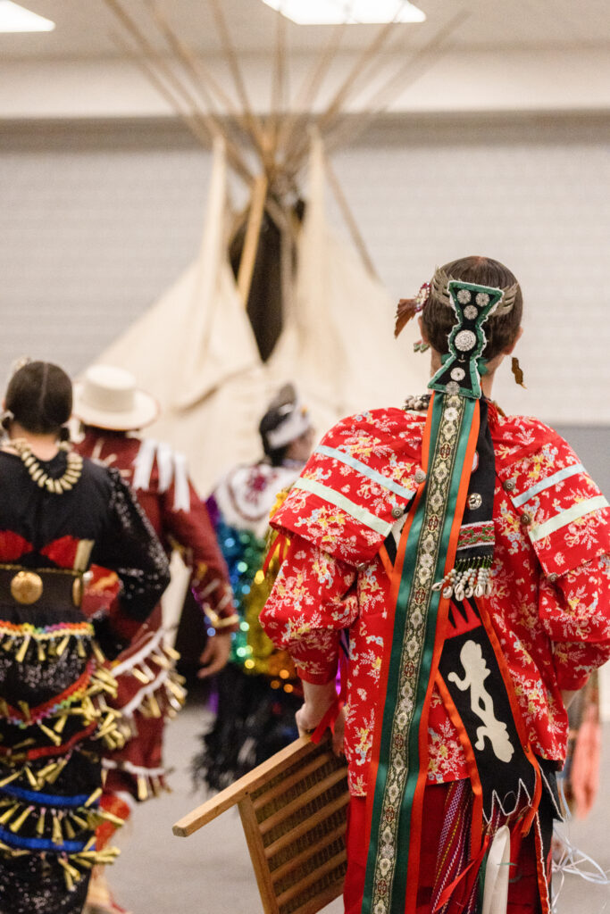 A group of dancers during a powwow dance. They have their backs turned, and are all wearing colorful regalia.
