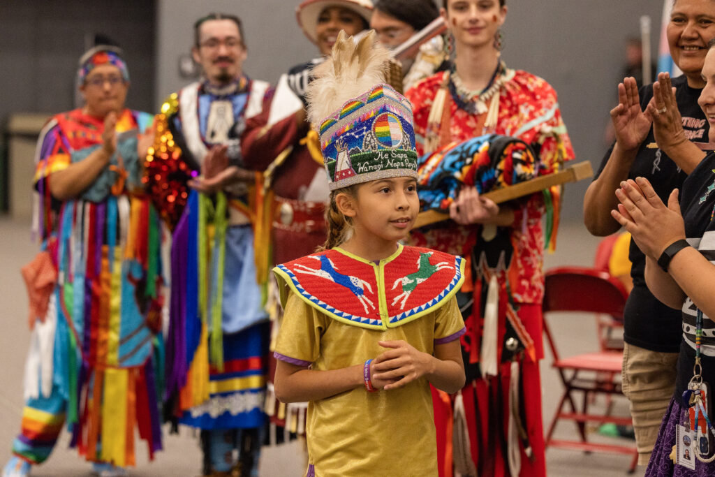 A child wearing a beaded collar and headdress standing in front of a crowd of people wearing colorful regalia and applauding.