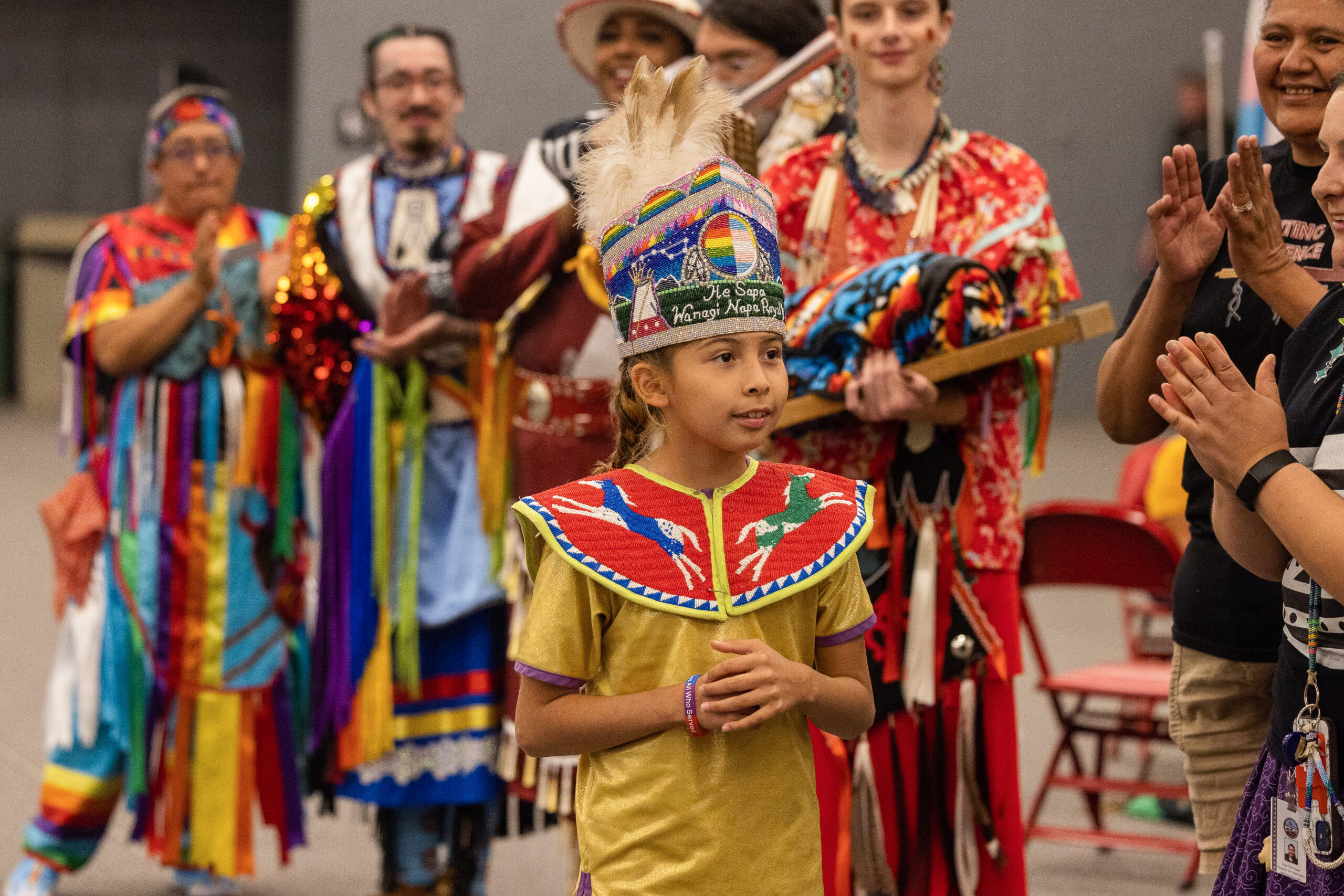 A child wearing a beaded collar and headdress standing in front of a crowd of people wearing colorful regalia and applauding.
